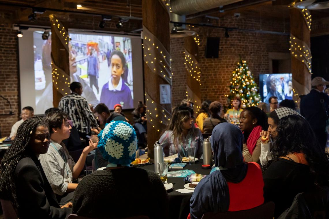 Group of people sat around tables with projector in background playing archive film