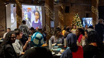 Group of people sat around tables with projector in background playing archive film