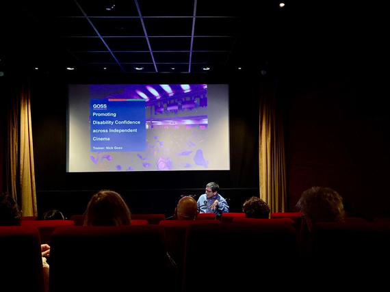 A man in a wheelchair sits in front of a cinema audience, screen says 'Nick Goss, Goss Consultancy'