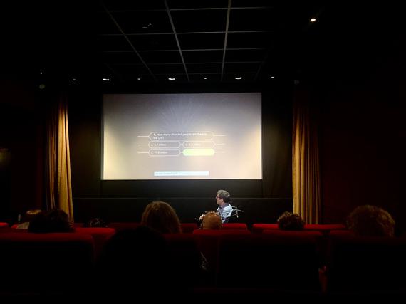 A man in a wheelchair sits in front of a cinema audience, screen says 'How many disabled peoiple are there in the UK?' with multiple choice answers - the correct answer, 14.1 million, is lit up in green.