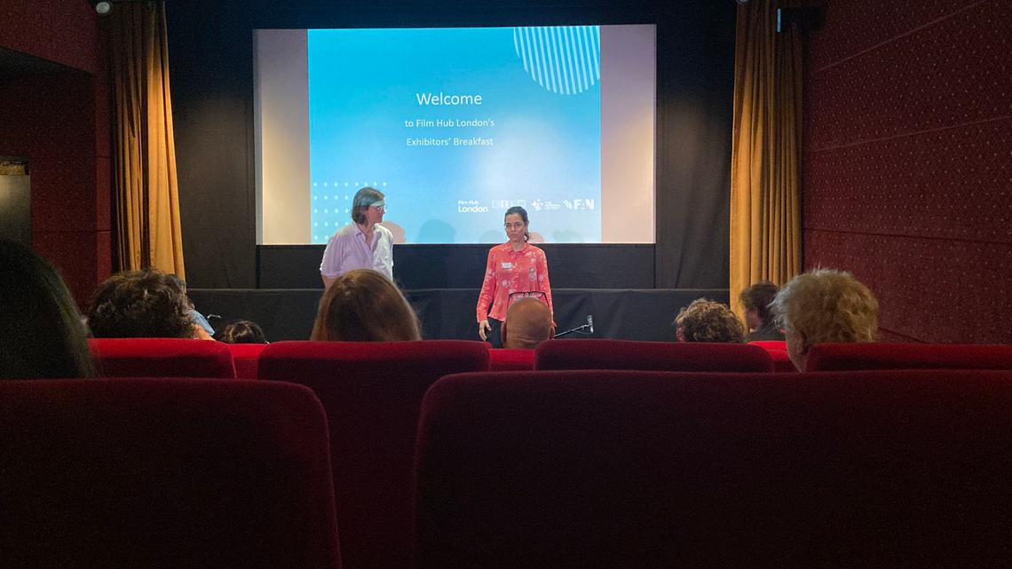 A man and woman stand in front of a cinema screen with a slide which says 'Welcometo Film Hub London’s Exhibitors’ Breakfast'