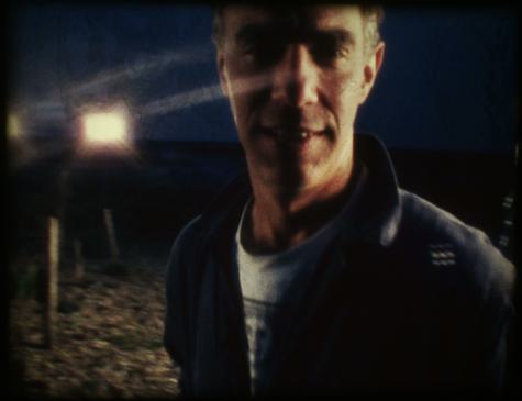 Derek Jarman looking at camera on the beach with a spotlight shining behind him.