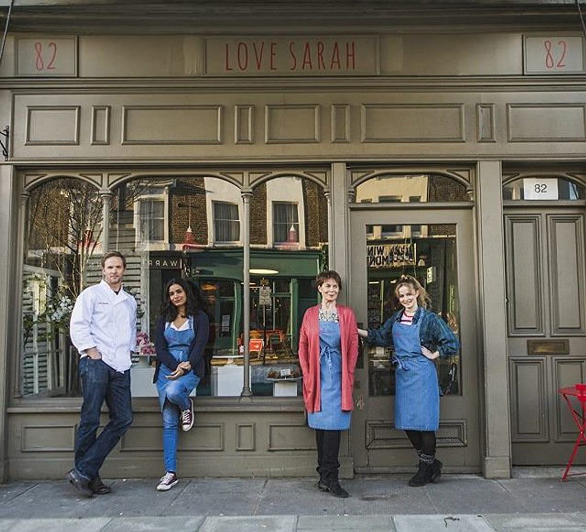 Still from Love Sarah. Four characters stand outside a bakery, looking directly at the camera