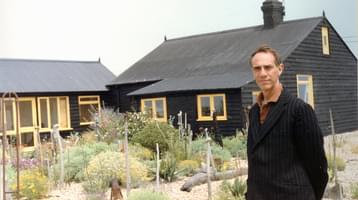 Derek Jarman standing in front of a house and garden, looking at the camera.