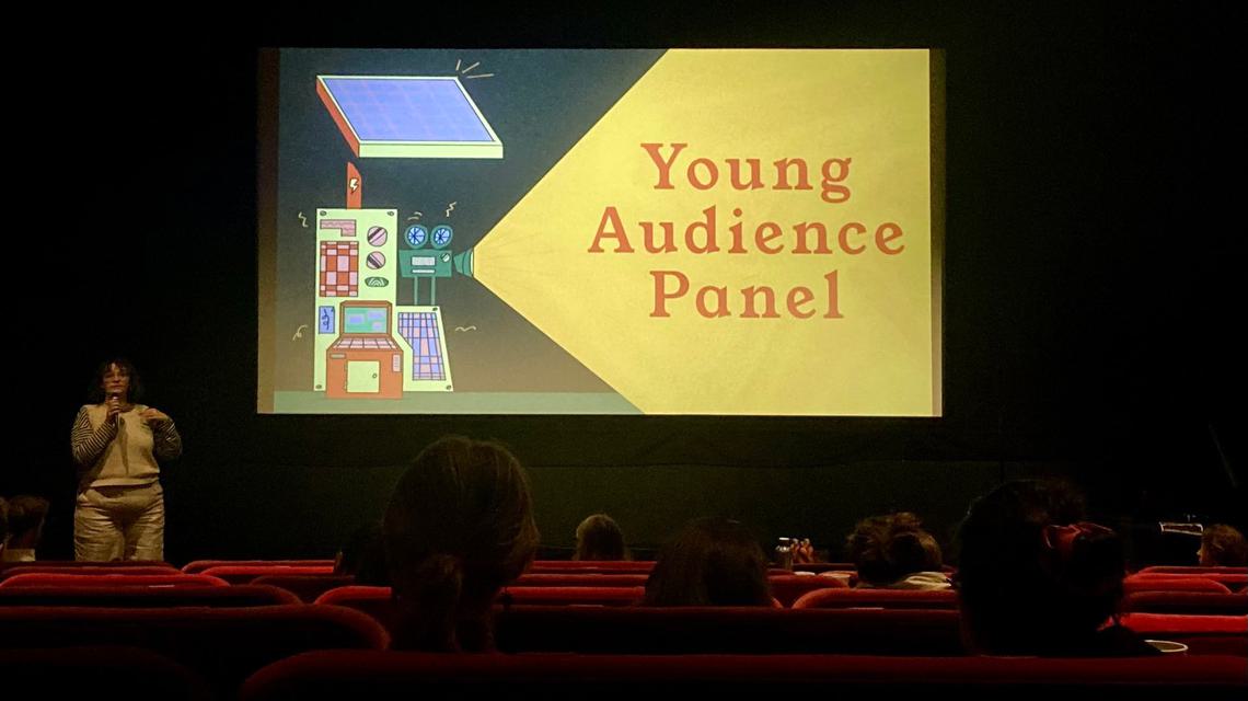 FAN Young Consultant Thea Berry addresses an audience in a cinema, in front of a screen with an illustration of a movie camera, and the words "Young Audience Panel"