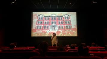FAN Young Consultant Thea Berry addresses an audience in a cinema, in front of a screen with an illustration of a large building